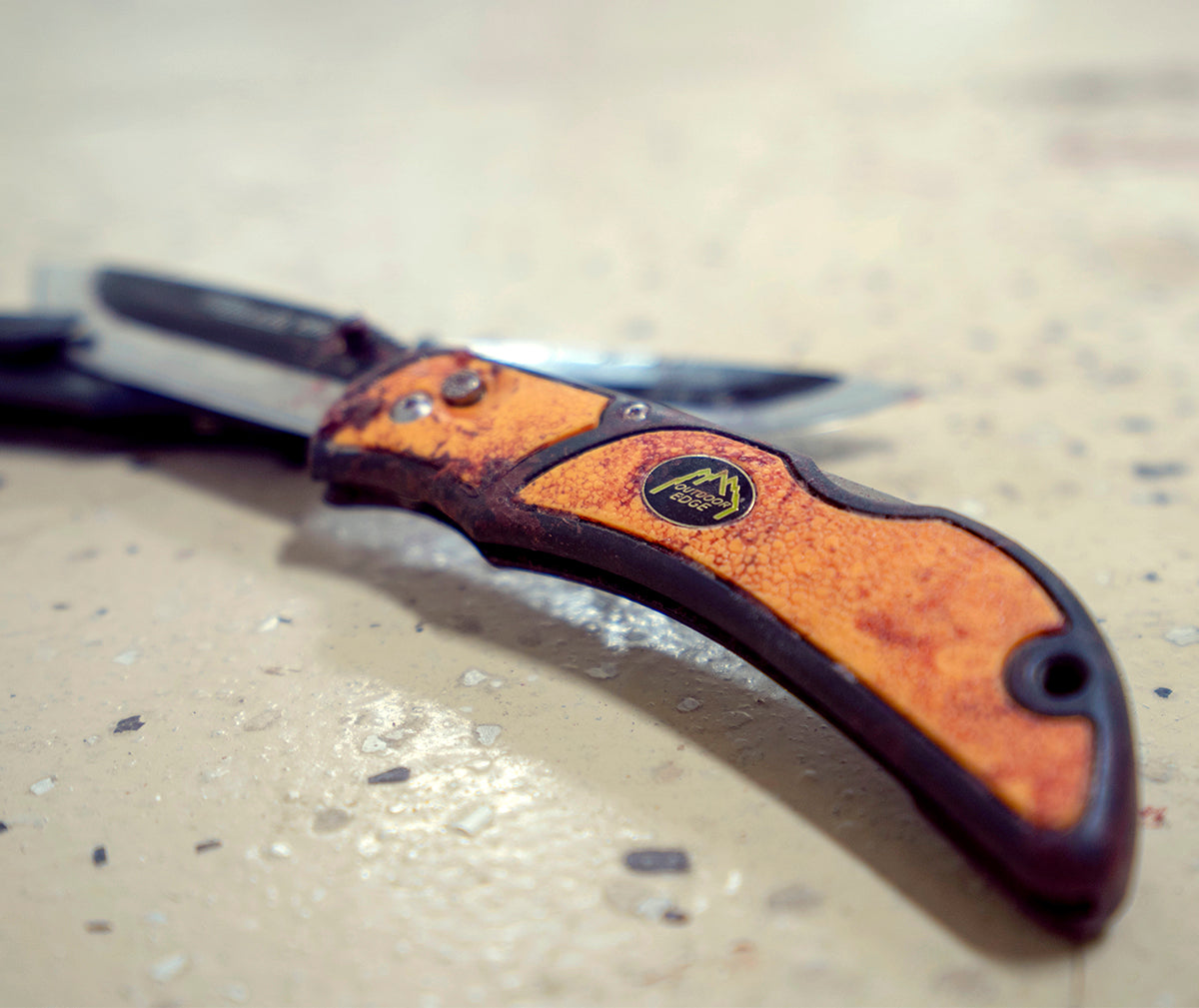 A close-up, artistic shot of a well-used, orange-handled Outdoor Edge folding knife resting on a flat surface.