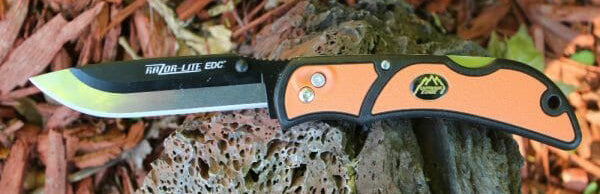 A close-up of the orange-handled Outdoor Edge 3.5" RazorLite EDC folding knife resting on a piece of wood outdoors. The knife features a replaceable blade system and a black pocket clip