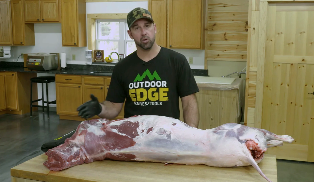 A man wearing an Outdoor Edge T-shirt stands in a kitchen behind a fully skinned deer carcass laid out on a large wooden cutting board, appearing to give instructions.
