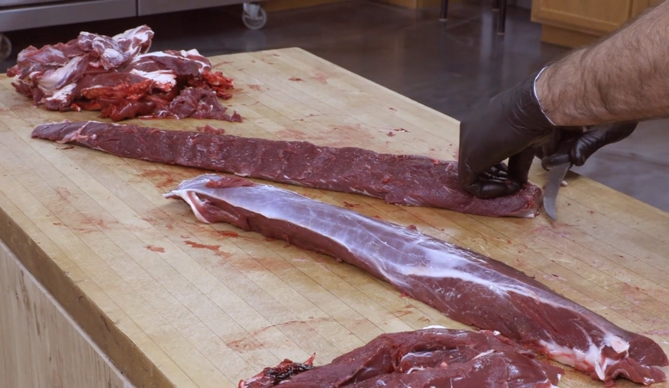 A close-up of a person wearing a black glove using a knife to trim the silverskin from two long pieces of venison backstrap on a wooden cutting board.
