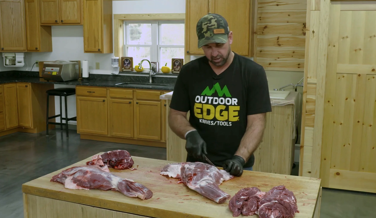 The man in the Outdoor Edge T-shirt processes a large cut of venison on a wooden cutting board in a kitchen setting.