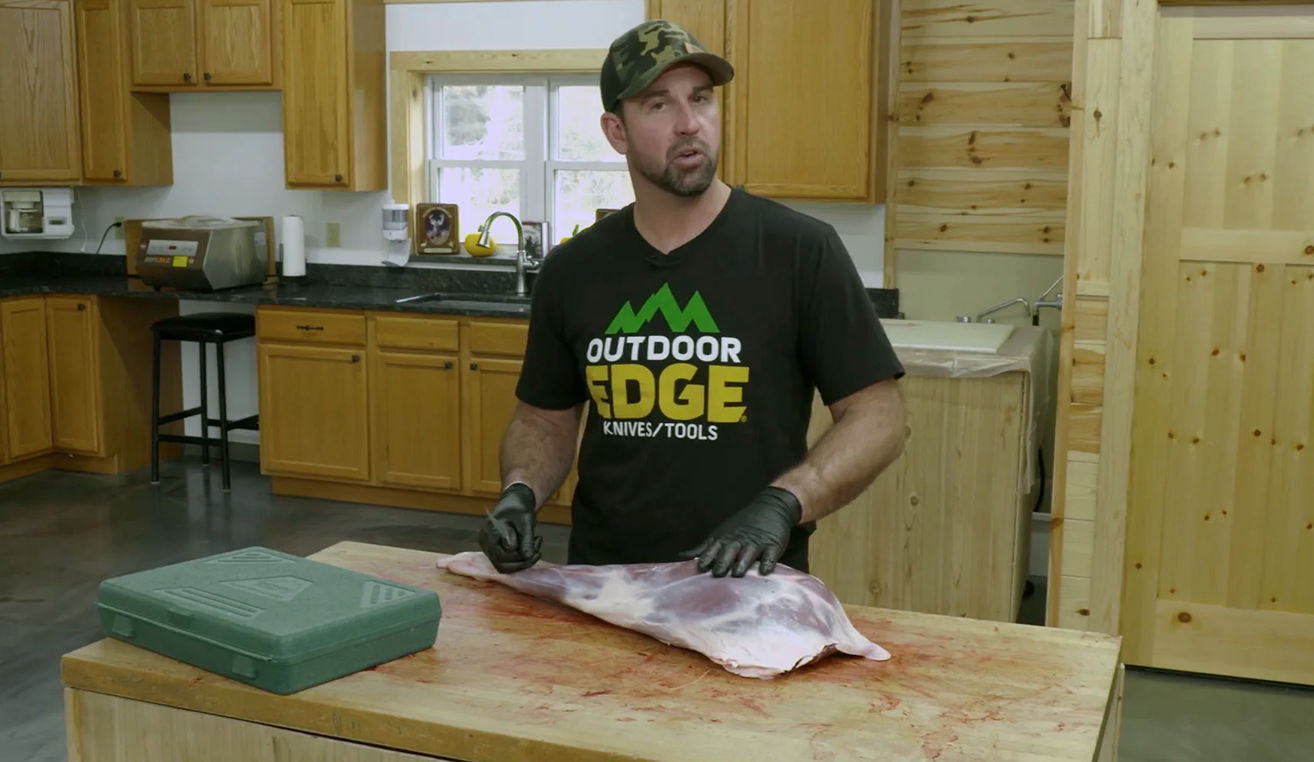 The man in the Outdoor Edge T-shirt stands over a large leg of venison on a cutting board, with a green Outdoor Edge processing kit case nearby