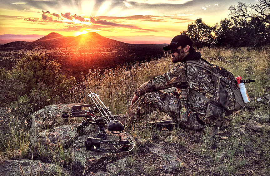 A hunter in full camouflage kneels on a rocky outcrop with his crossbow, looking out at a mountain range during a vibrant sunset.