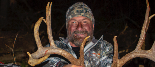 A close-up photo of a smiling hunter in camouflage holding up the large antlers of a harvested buck at night.