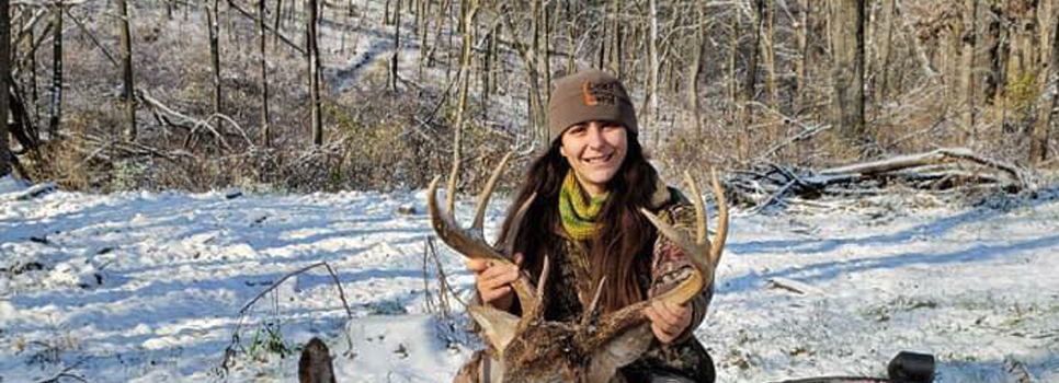 A hunter in camouflage kneels in the woods at dusk beside a harvested buck, with an Outdoor Edge SwingBlade and folding saw from the SwingBlaze Pak placed on the deer