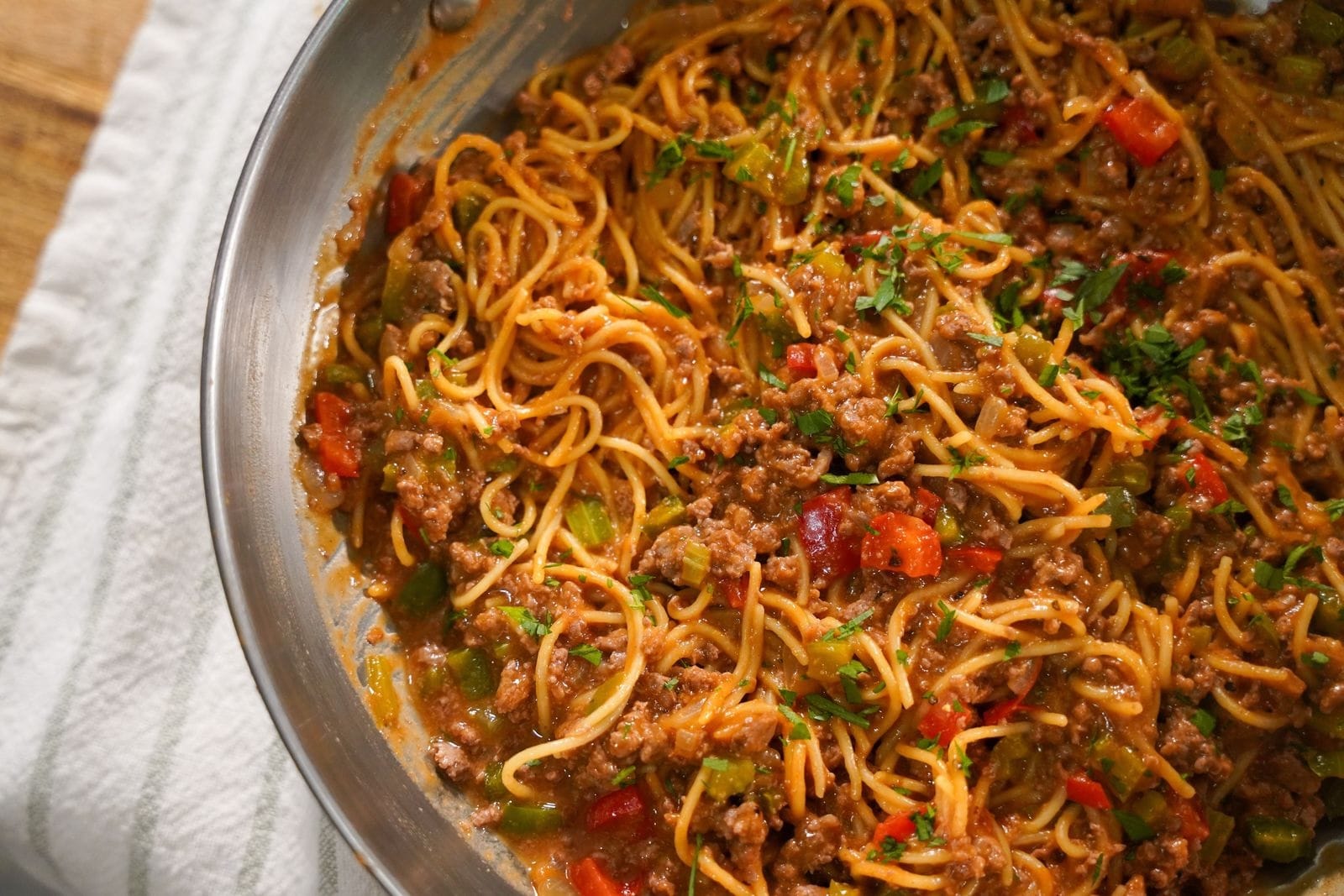 Overhead view of dirty venison spaghetti in a stainless steel skillet with ground venison, red and green bell peppers, and fresh parsley in a rich Cajun tomato sauce