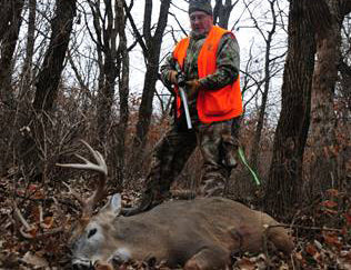 A hunter wearing camouflage and a blaze orange safety vest stands in the woods with his foot on a large harvested whitetail buck.