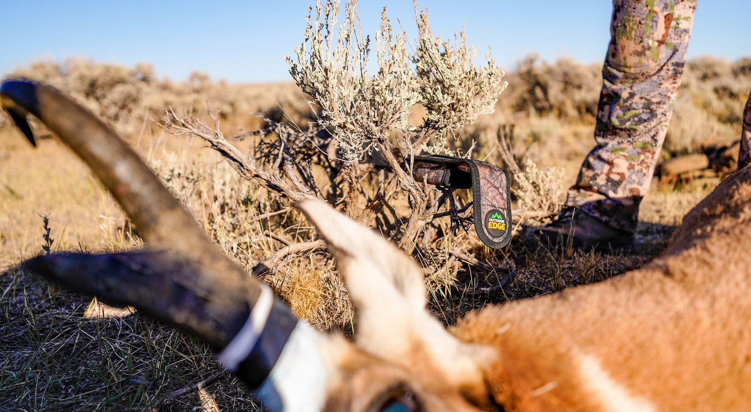 A hunter in camouflage stands in a dry, grassy field next to a freshly hunted antelope with distinct horns.