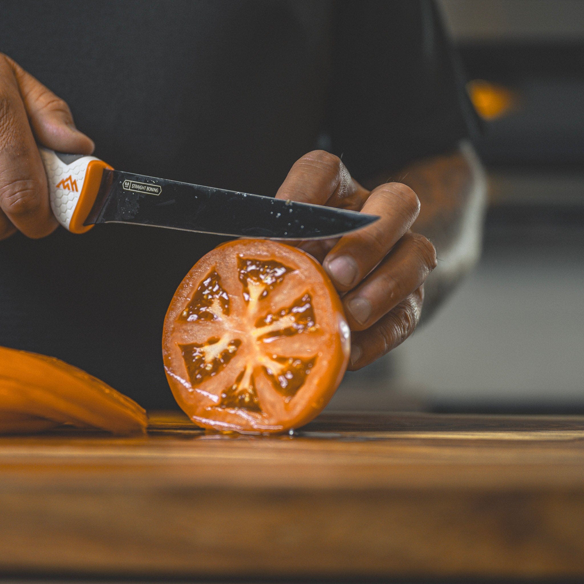 Hand holding a knife cutting a tomato on a wooden surface