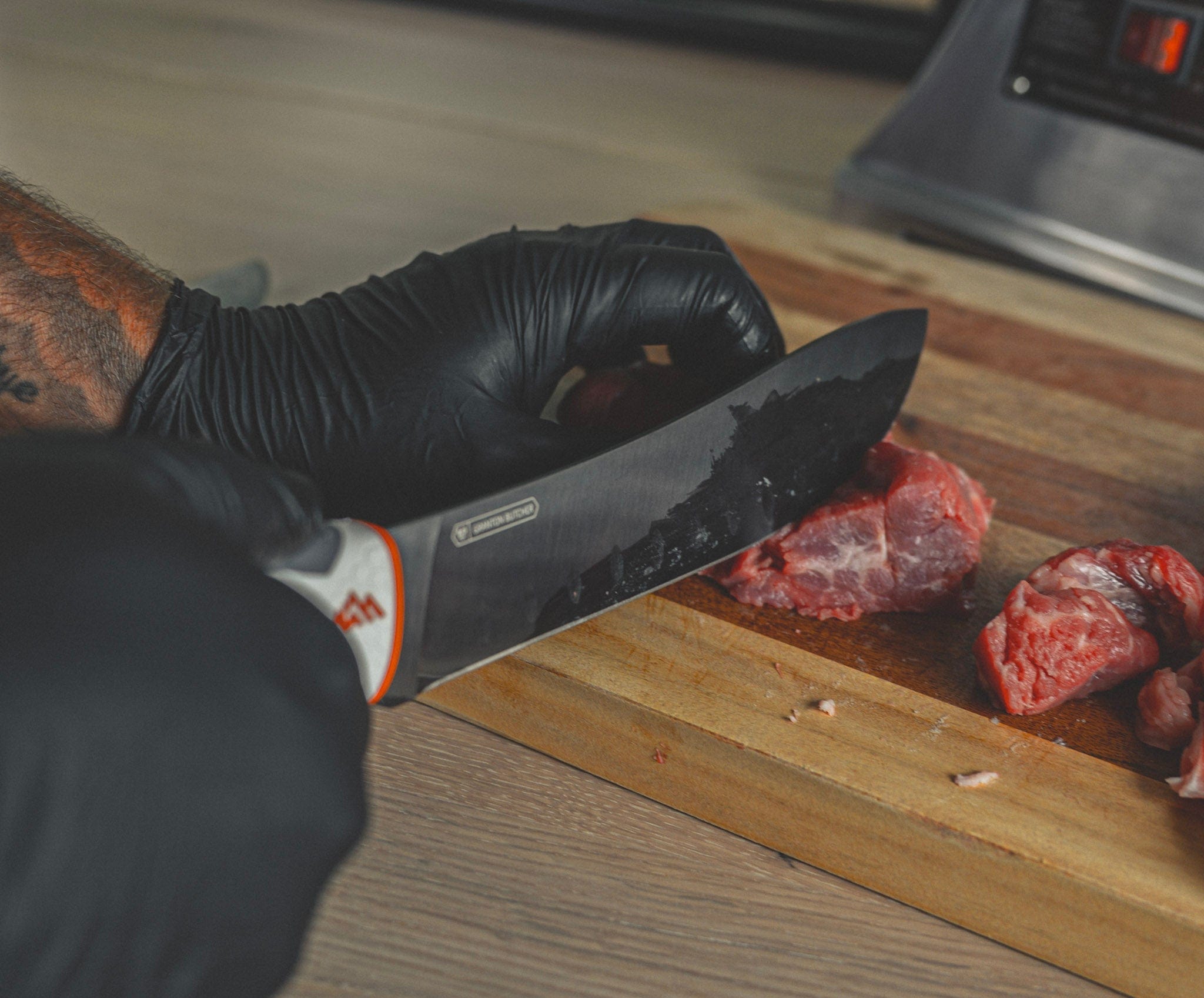 Person cutting meat on a wooden board with a knife, wearing black gloves.
