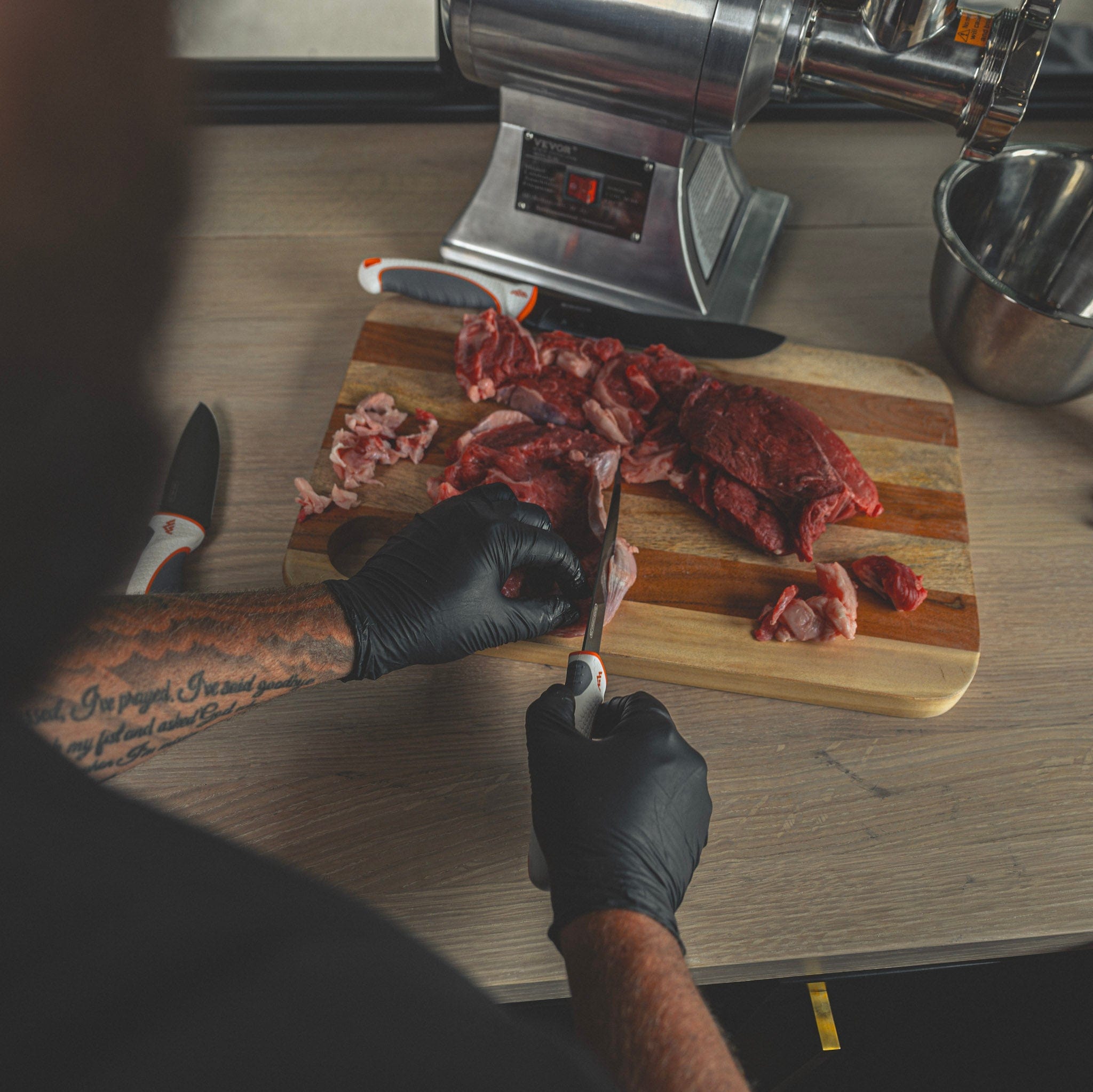 Person cutting raw meat on a wooden cutting board with a meat grinder in the background.