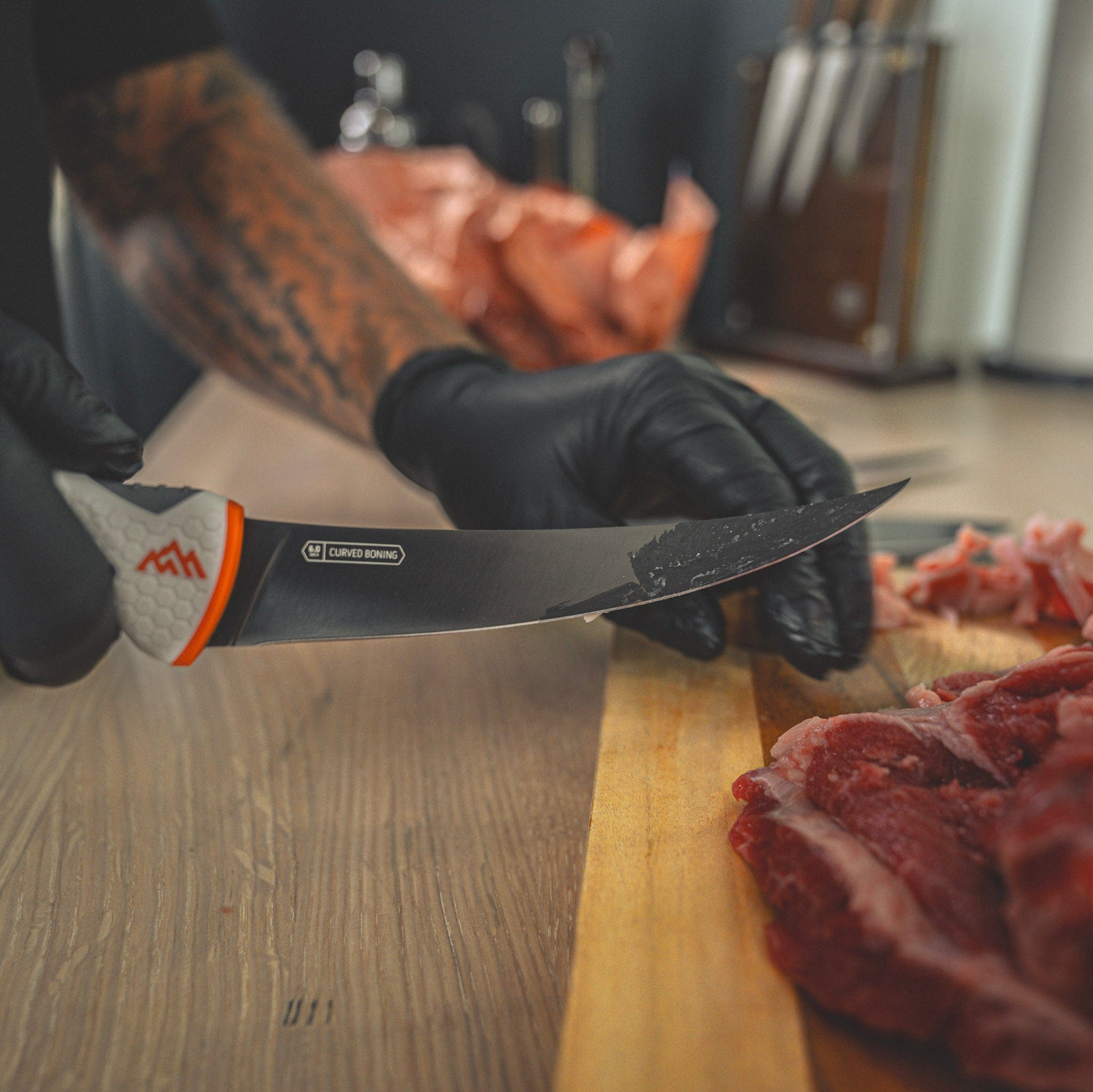 Person wearing black gloves and a gray glove with orange accents holding a knife over raw meat on a wooden cutting board.
