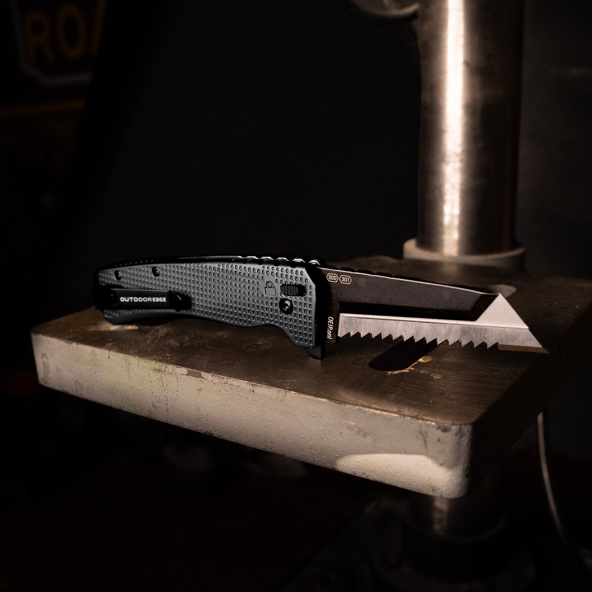 "An Outdoor Edge knife with a serrated tanto blade and grey G10 handle resting on a metal block in a workshop."