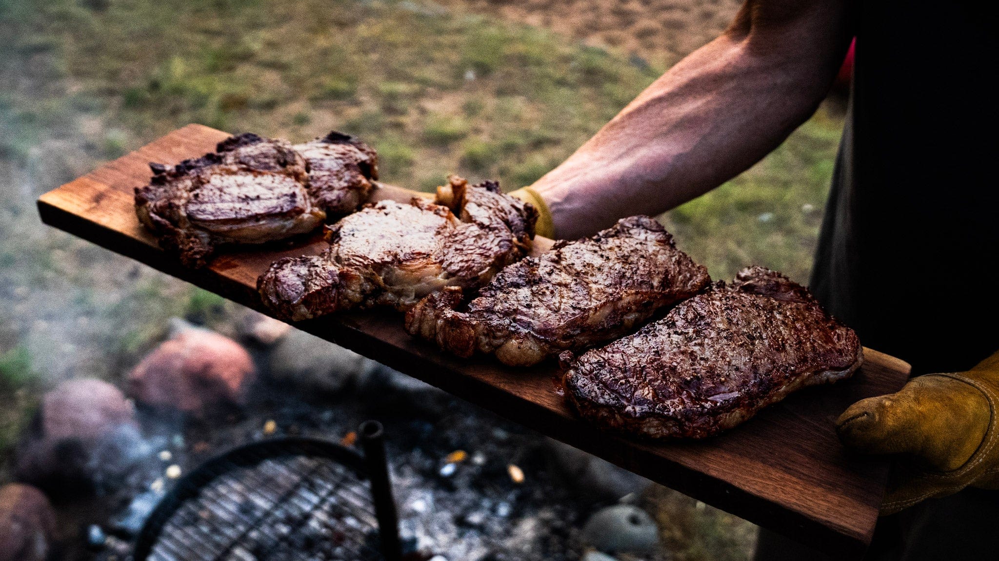 Person holding a wooden board with grilled steaks outdoors