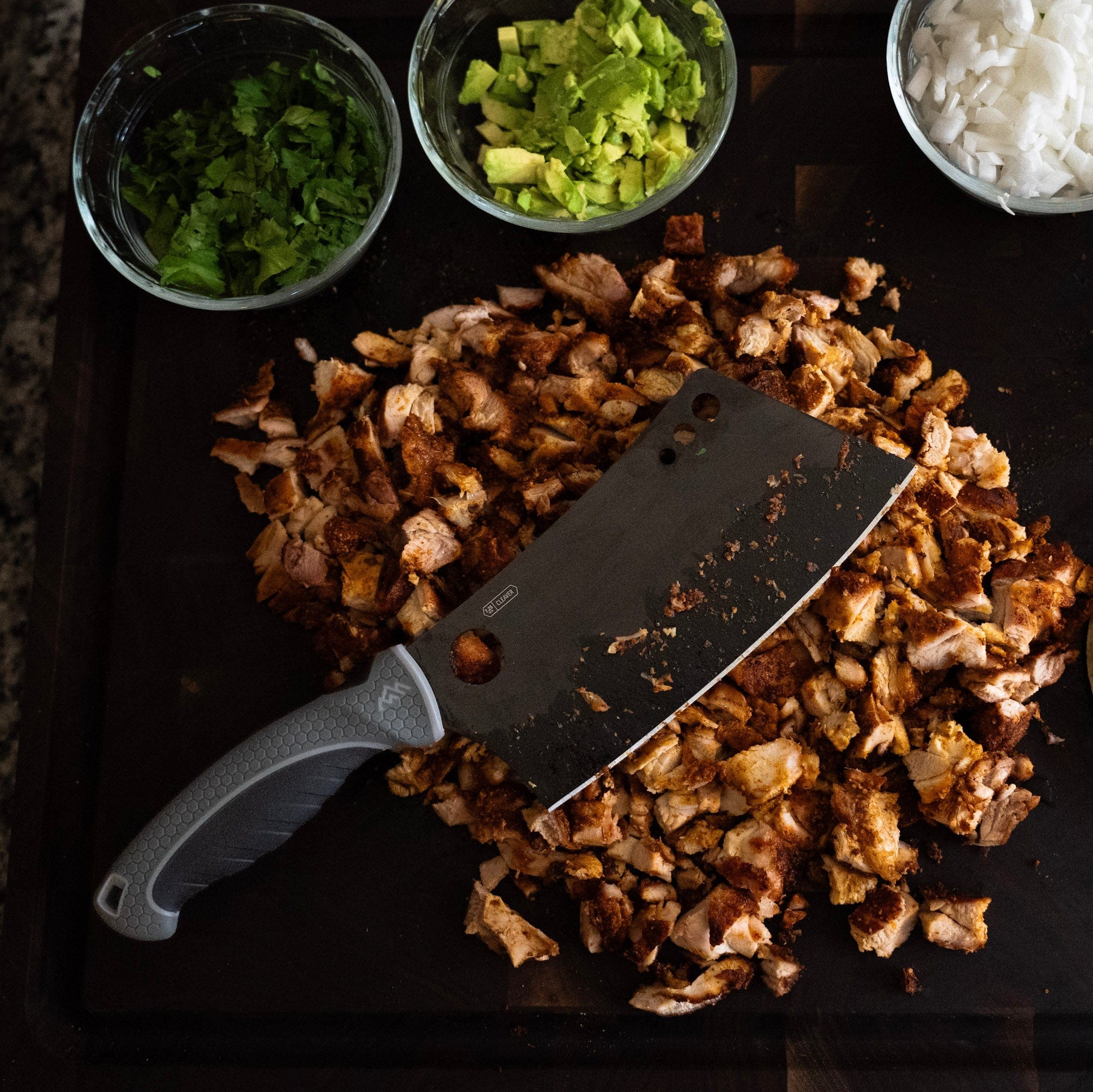 Chopped meat on a cutting board with a knife, surrounded by small bowls of chopped vegetables.