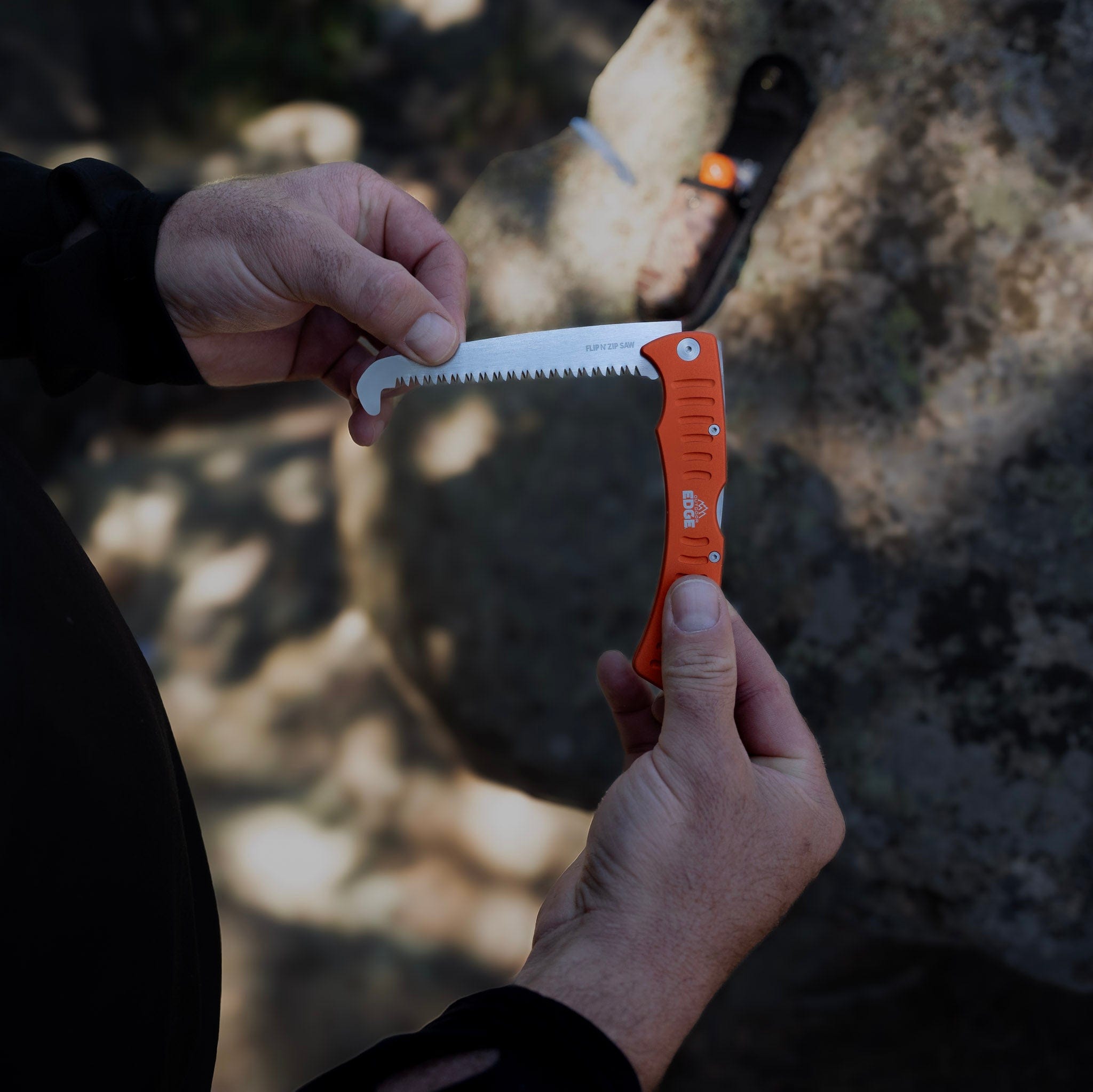 Person holding an orange folding saw with a blurred natural background