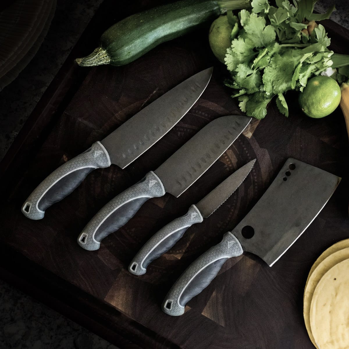 4 kitchen knives sitting on a dark wood cutting board with fresh green produce