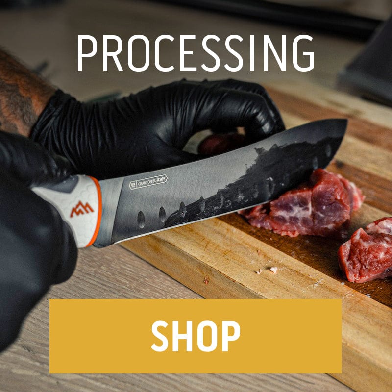 A person wearing black nitrile gloves uses a large Outdoor Edge processing knife to slice a raw steak on a wooden cutting board."