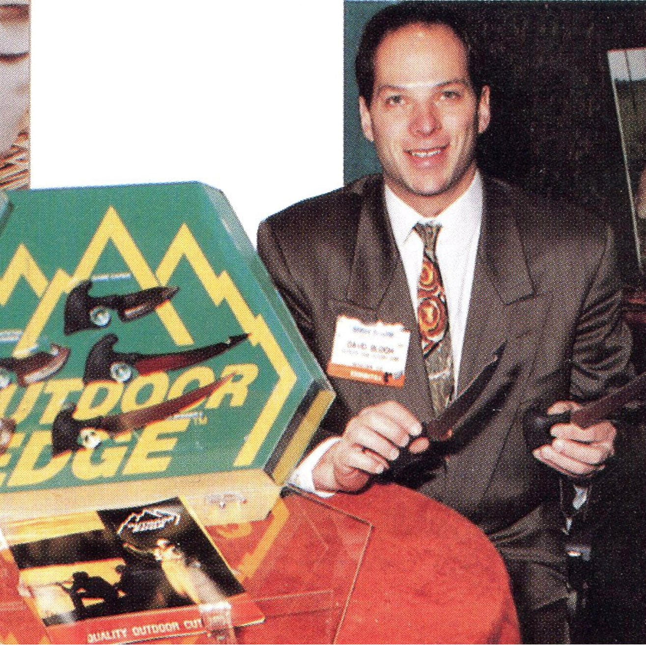 "A vintage photo of Outdoor Edge founder David Bloch at a trade show, holding a knife next to a product display."