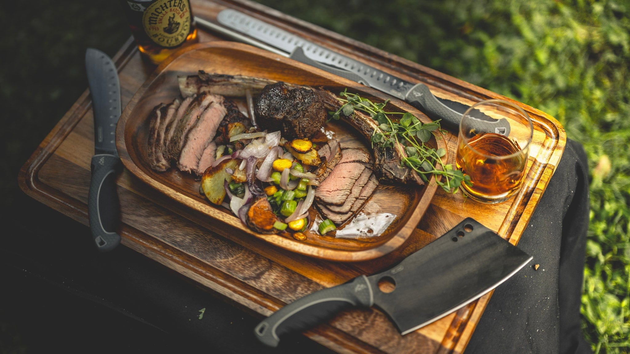 Wooden cutting board with sliced meat, vegetables, and a bottle of beer on a grassy background