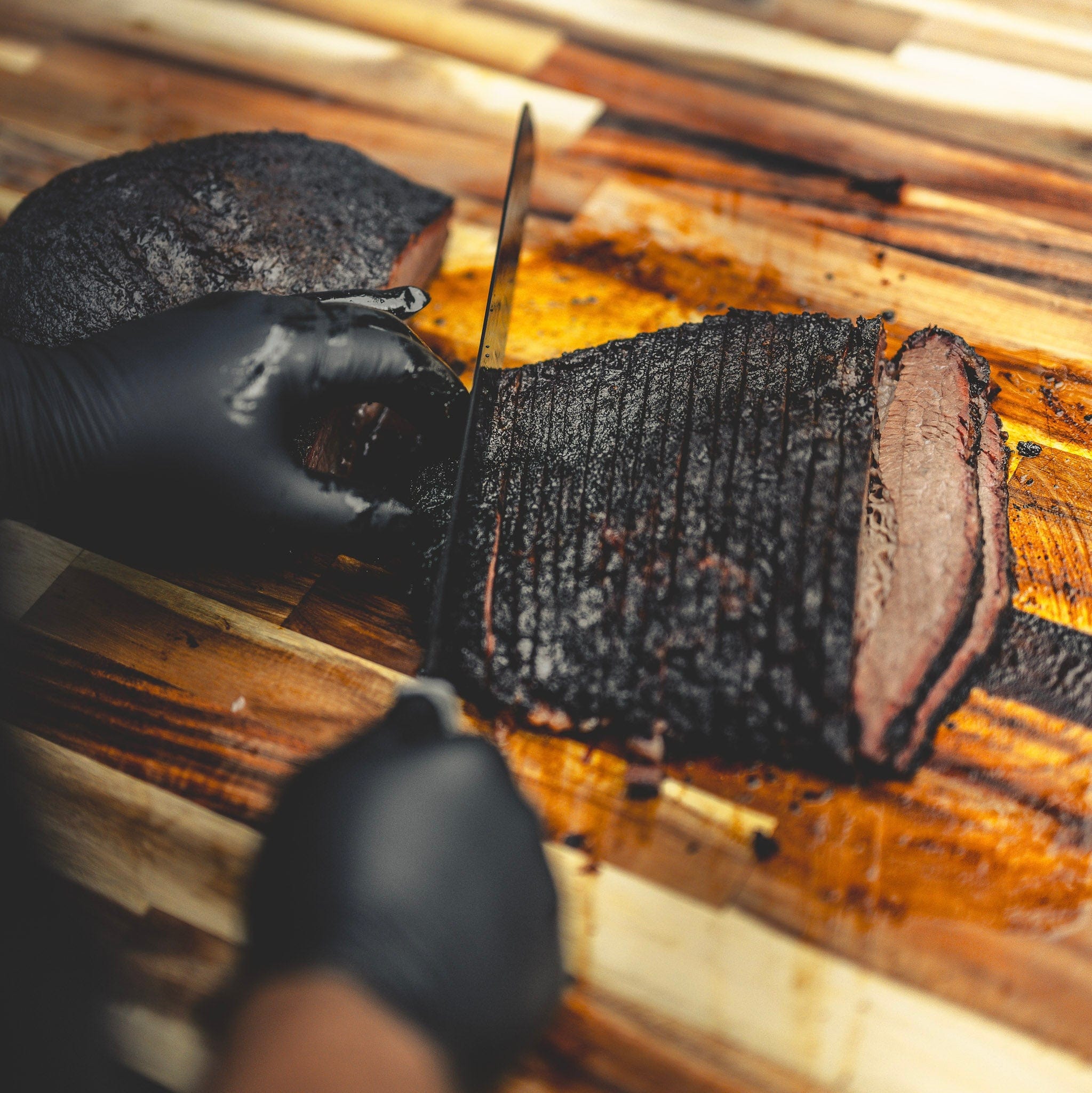 Person wearing black gloves slicing a brisket on a wooden cutting board.