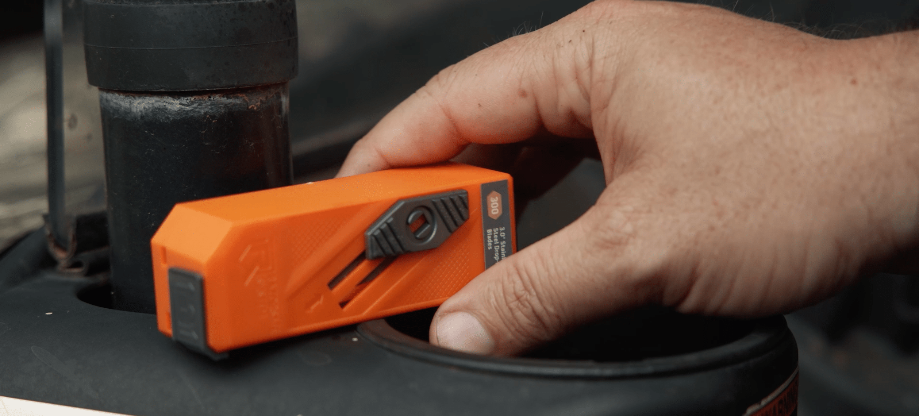 A hand with dirt under its fingernails is holding a small, bright orange and black tool.