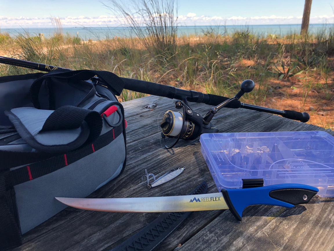 An Outdoor Edge ReelFlex fillet knife lies on a wooden picnic table next to a fishing rod and reel, tackle bag, and tackle box, with a beach scene in the background