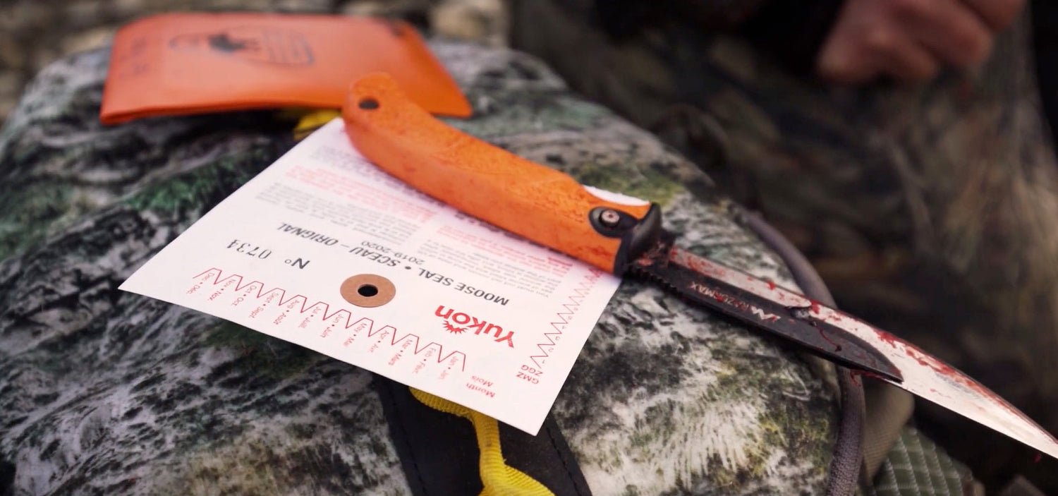  A close-up of an orange-handled Outdoor Edge knife with a Yukon hunting tag attached, resting on a camouflage surface.