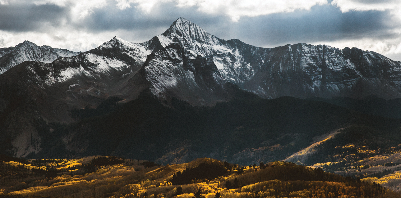 A dramatic mountain landscape showing sharp, snow-dusted peaks rising above a dark, forested valley under a cloudy sky. 