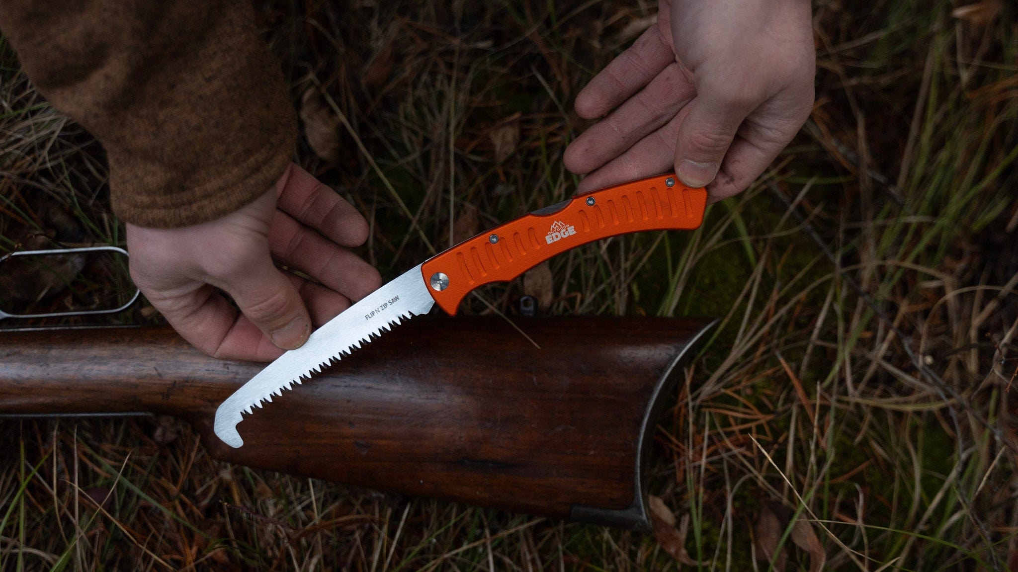 Person using an orange folding knife with a saw blade on a wooden handle outdoors.