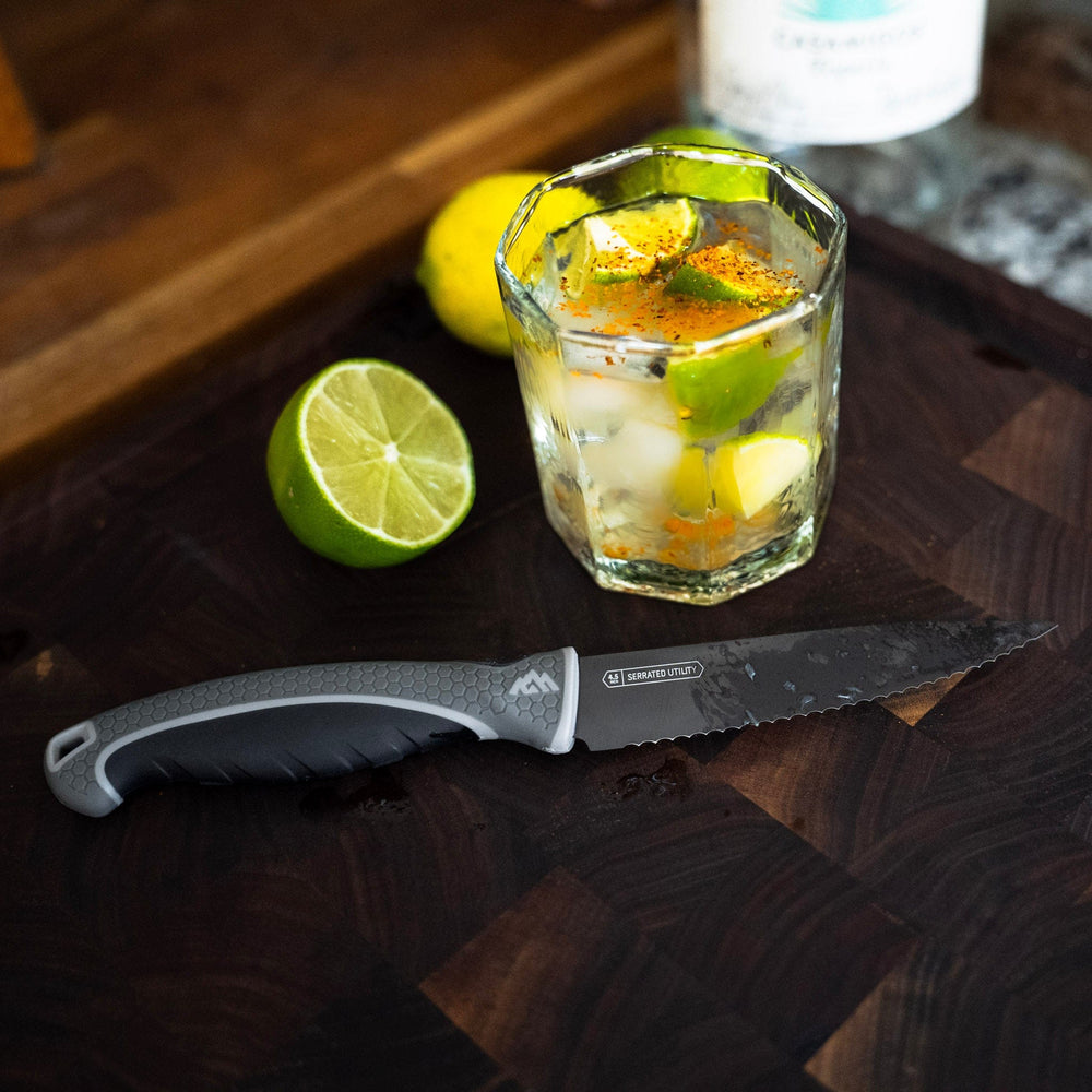 Knife on a wooden surface with a glass of lime and orange slices, and a bottle in the background.