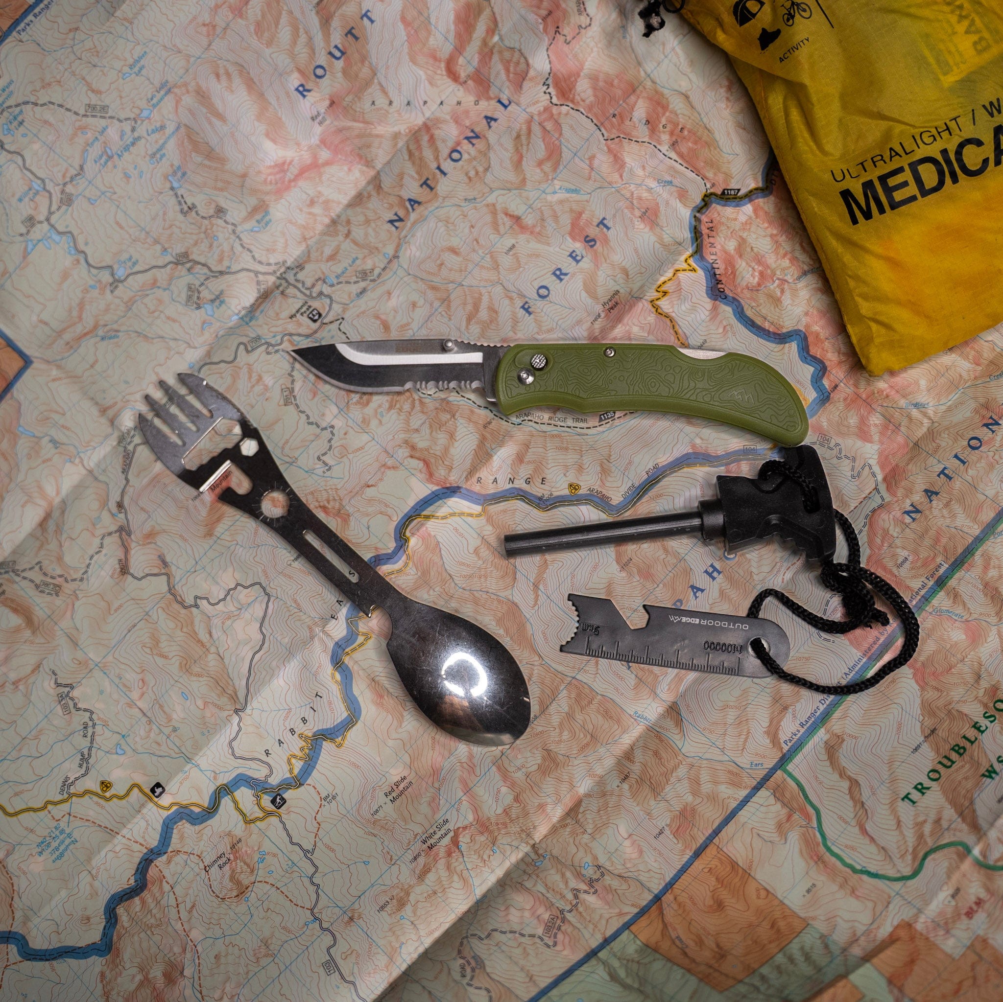 Fork, knife, and spoon set on a map with a fire starter and medical bag in the background.