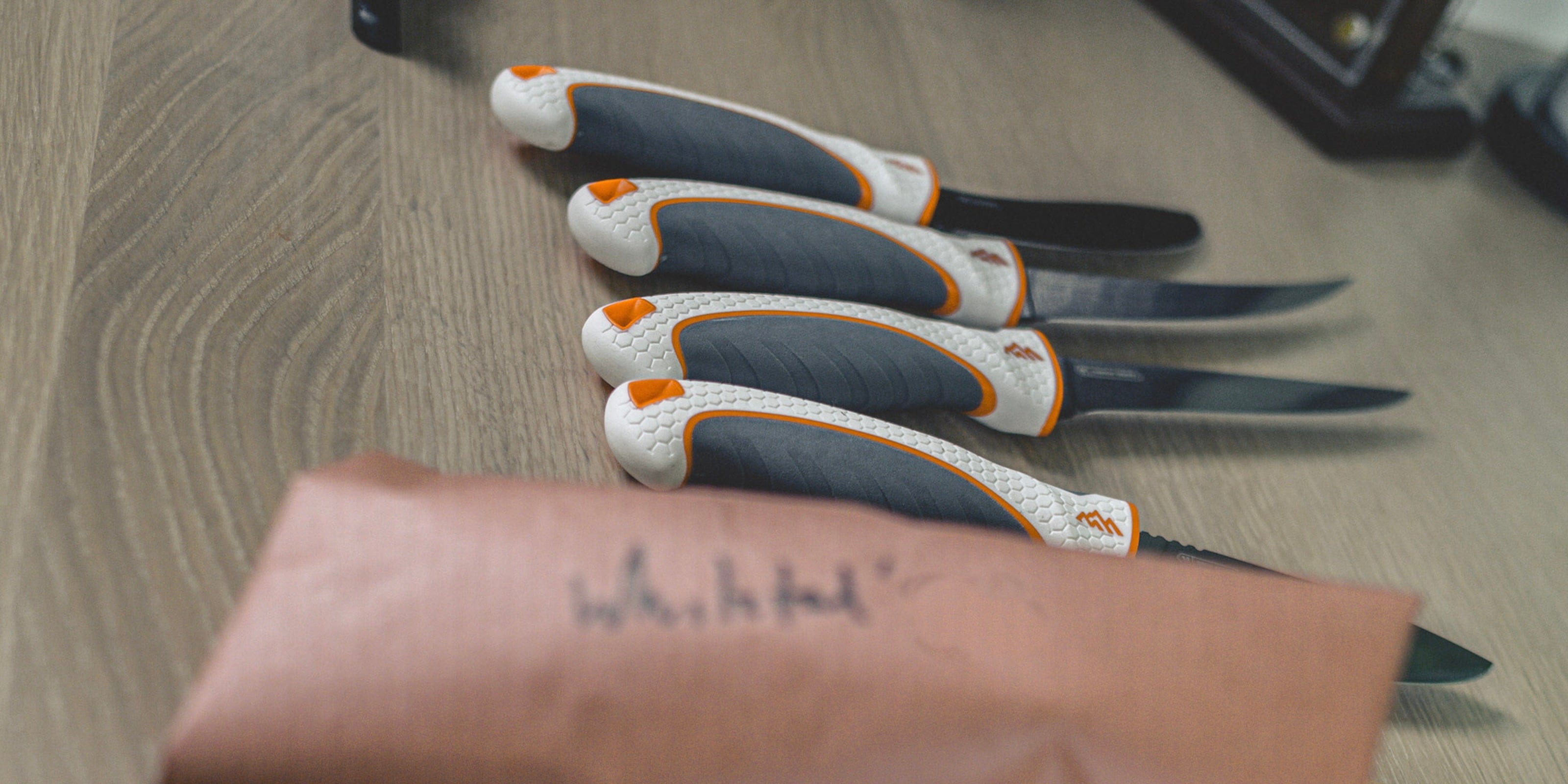 Set of four pocket knives with orange accents on a wooden surface, with a brown leather sheath in the foreground.