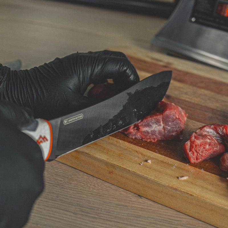 Person in black gloves cutting meat on a wooden board with a knife.